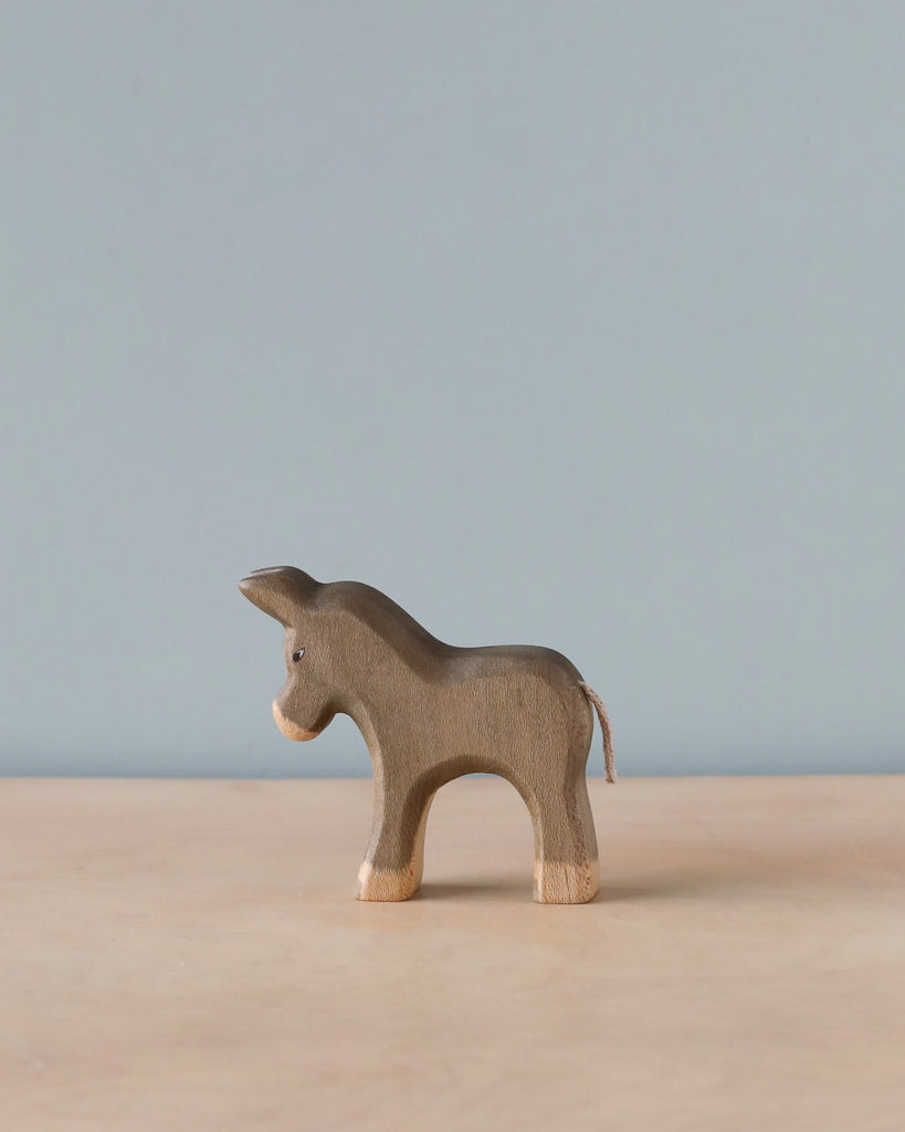 A Handmade Holzwald Small Donkey stands on a plain surface against a light blue backdrop, featuring textured wood and visible grain, with a contrasting smooth, white tail.