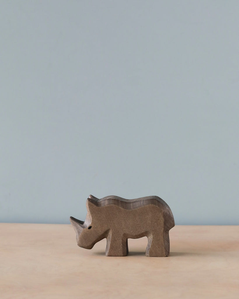 A small Handmade Holzwald Baby Rhino figurine, crafted from high-quality sustainable materials, stands on a smooth surface against a plain light blue background, showcasing its simplistic and artistic carved details.