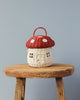 An Olli Ella Red Mushroom Basket, shaped like a mushroom with a red cap and white stalk, placed on a wooden stool against a neutral gray background.