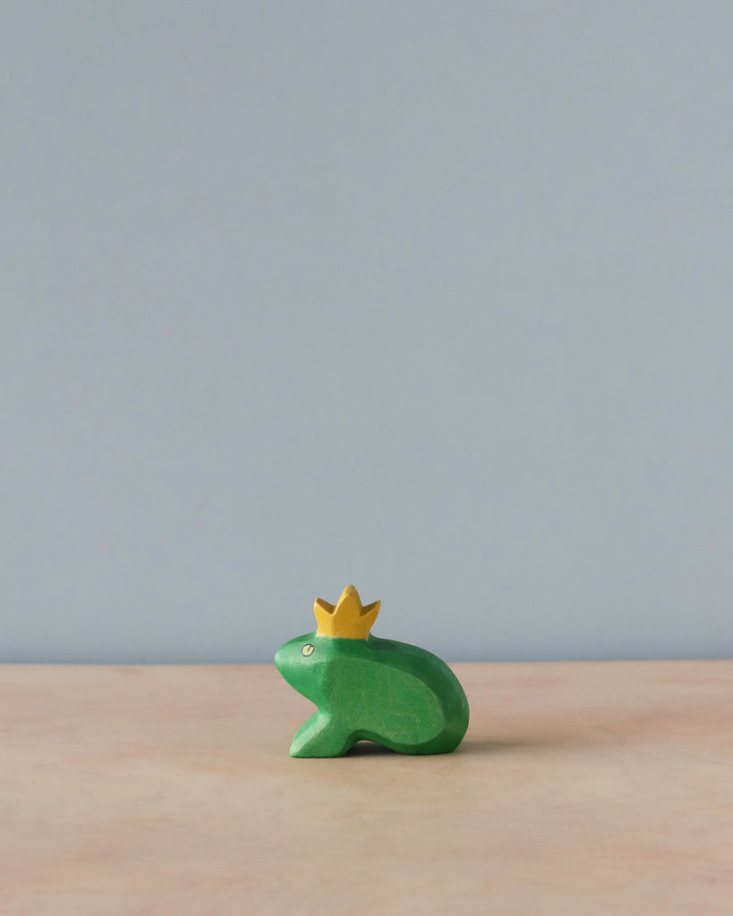 A small, green, wooden Handmade Holzwald Frog King figurine wearing a yellow crown, placed on a wooden surface against a plain, light blue background. Made from sustainable toys materials.