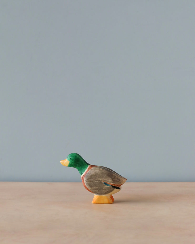 A small, high-quality Handmade Holzwald Male Duck figurine with a green head, grey body, and orange feet, standing against a plain, light blue background.