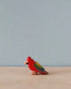 A high-quality Handmade Holzwald Parrot Bird toy with red, yellow, and green feathers, positioned on a plain gray surface against a light blue background.
