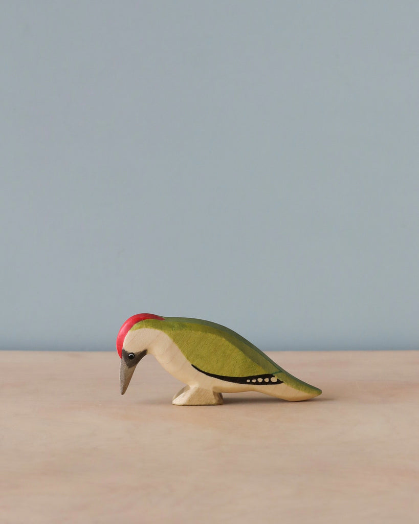 A Handmade Holzwald Woodpecker Bird with a red head, green body, and yellow and black details stands against a simple blue background with a light brown surface. This high-quality wood toy blends craftsmanship and