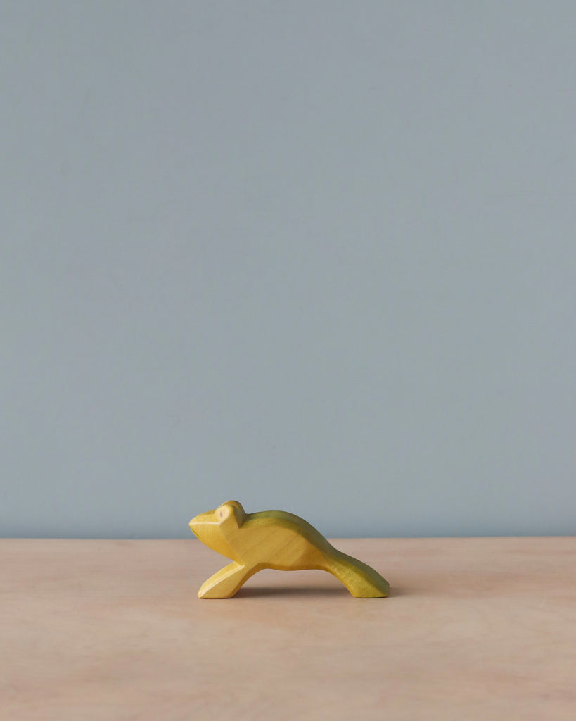 A small, carved Handmade Holzwald Jumping Frog figurine is positioned on a smooth wood surface against a gentle blue background. The figurine showcases visible grain, enhancing its rustic appearance.