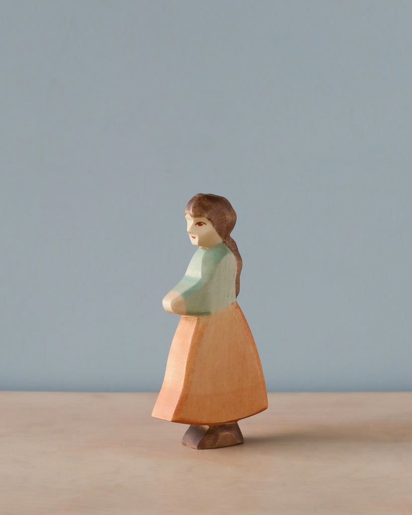 A Handmade Holzwald Goose Girl figurine of a woman with a subtle smile, painted in soft pastel colors using natural dyes, stands against a plain light blue background. The figurine has a pink skirt, green.
