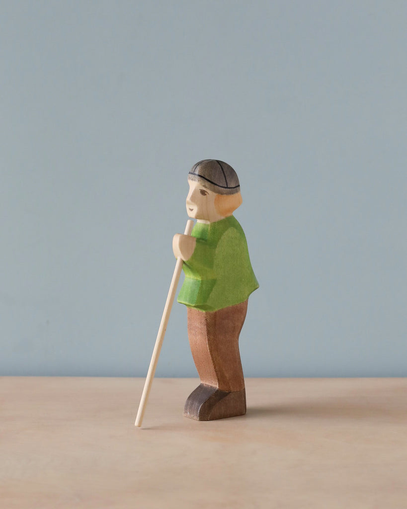 A handmade Holzwald Shepherd figurine of an elderly man with a cane, wearing a cap and green shirt, standing against a pale blue background.
