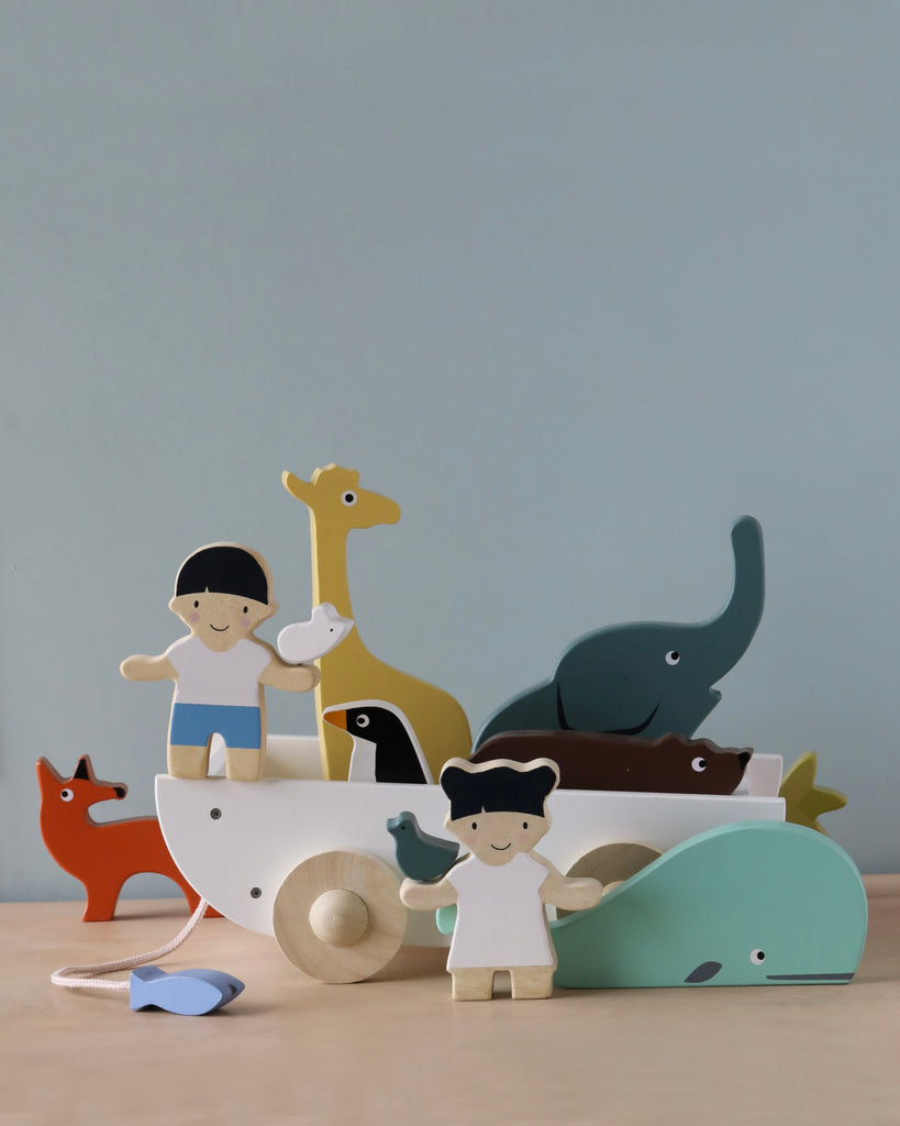 A colorful collection of wooden toys including animals and dolls on a gray surface against a light background. The Friend Ship Pull Toy, made from sustainable rubber wood, carries smaller figures and animals.