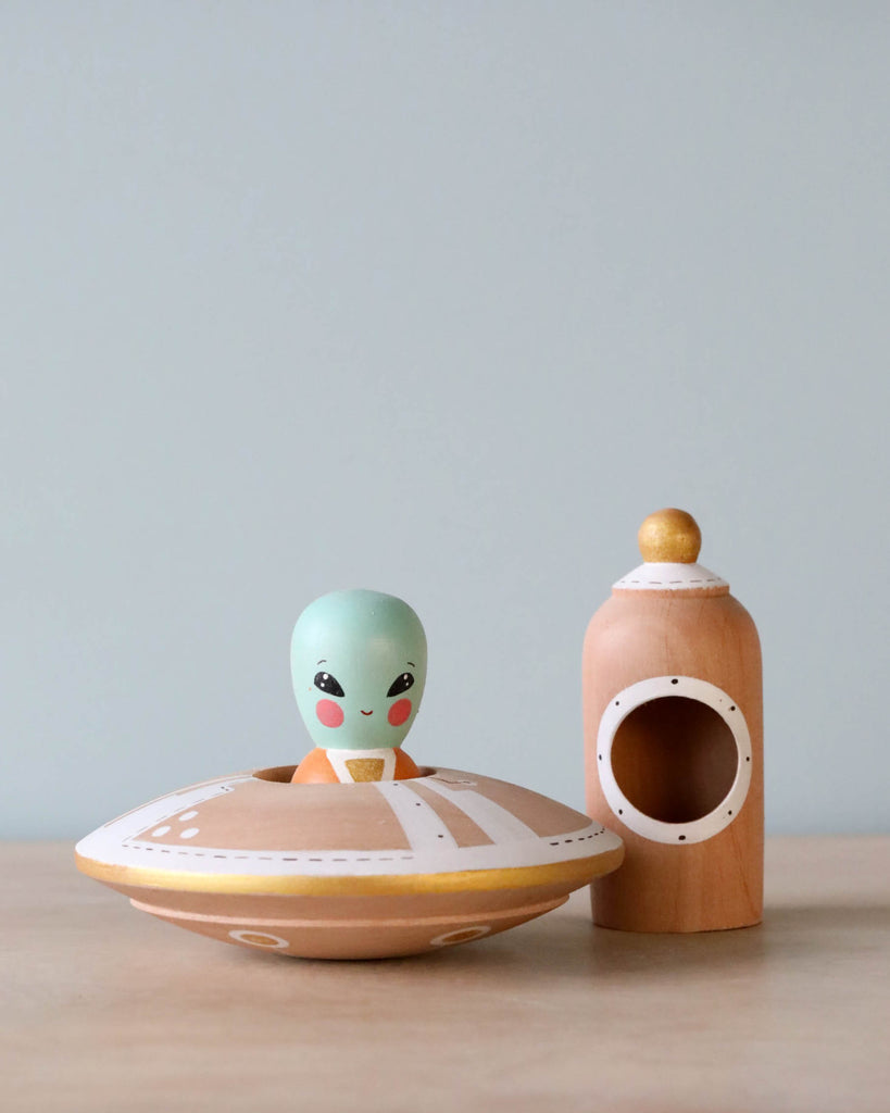 A small, cute Handmade Wooden UFO With Alien, resembling a hand-painted alien, sits in a handmade UFO-shaped object next to an empty wooden birdhouse on a plain background.