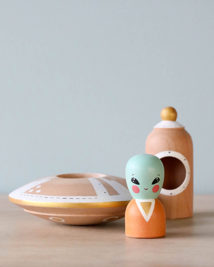 A Handmade Wooden UFO With Alien toy with a removable dome paired with a small hand-painted wooden alien figure, both resting on a wooden surface against a light blue background.