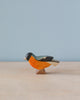 A Handmade Holzwald Bullfinch Bird figurine stands on a light wood surface against a pale blue background, featuring bright orange, yellow, and black colors on its body. This high-quality wooden toy is crafted to delight.