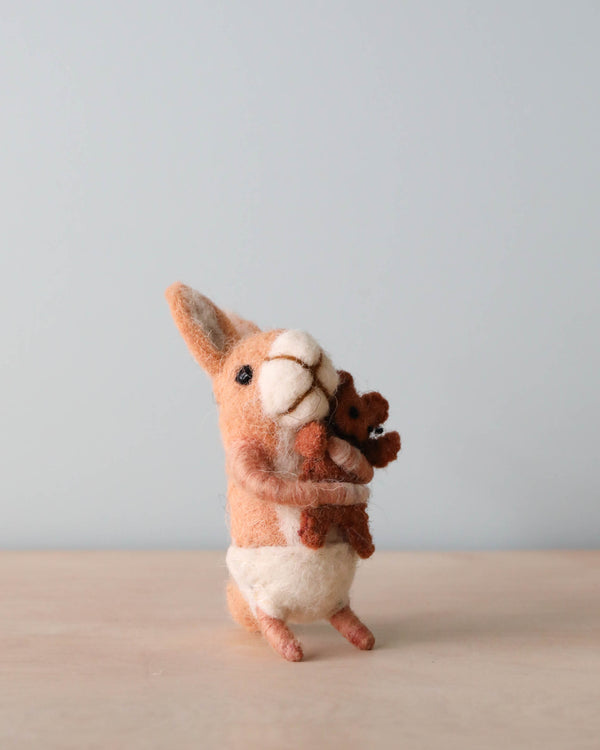 A hand felted sculpture of a Felt Bunny With Teddy Bear holding a joey, positioned against a plain, light-blue background. The kangaroo stands upright, featuring detailed texture and natural colors. Made in Nepal.