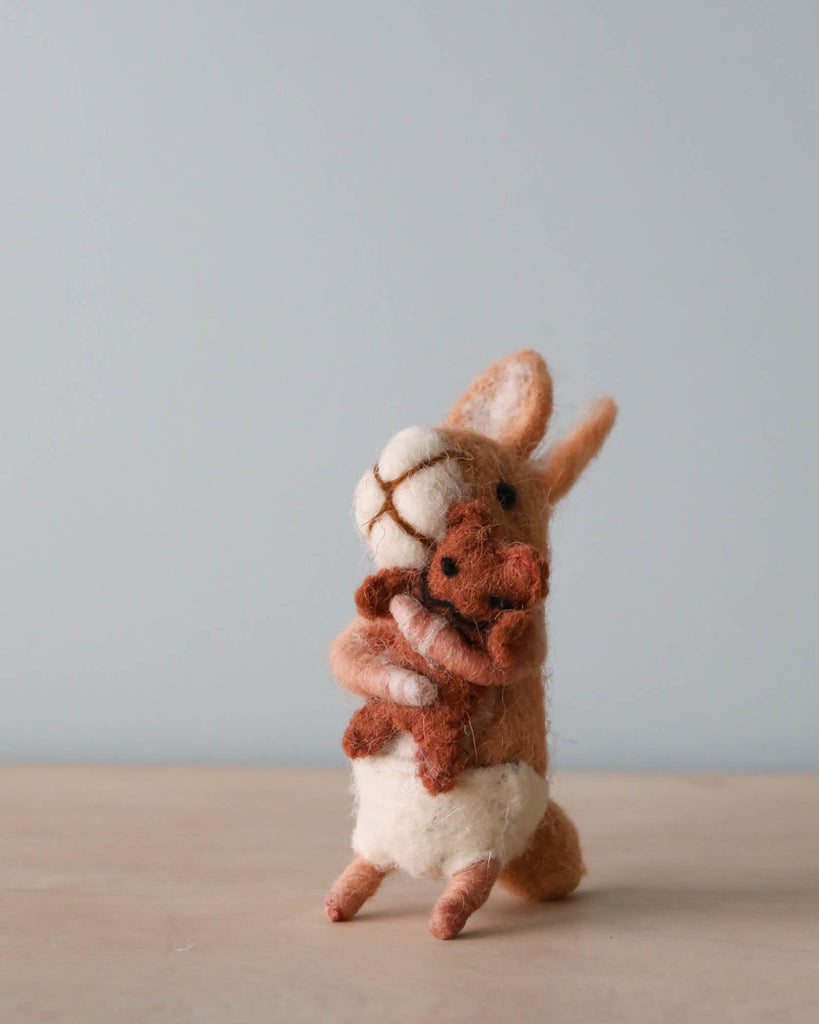 A hand-felted sculpture of a Felt Bunny With Teddy Bear standing upright, hugging a smaller rabbit, set against a plain light blue background.