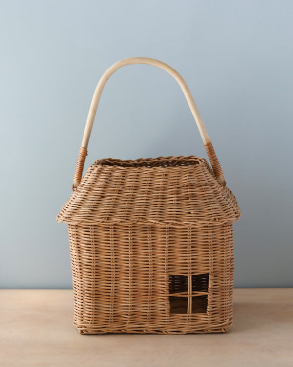 A handmade Olli Ella Rattan Hutch Basket - Large shaped like a small house with a pitched roof and a curved handle, set against a plain blue background.