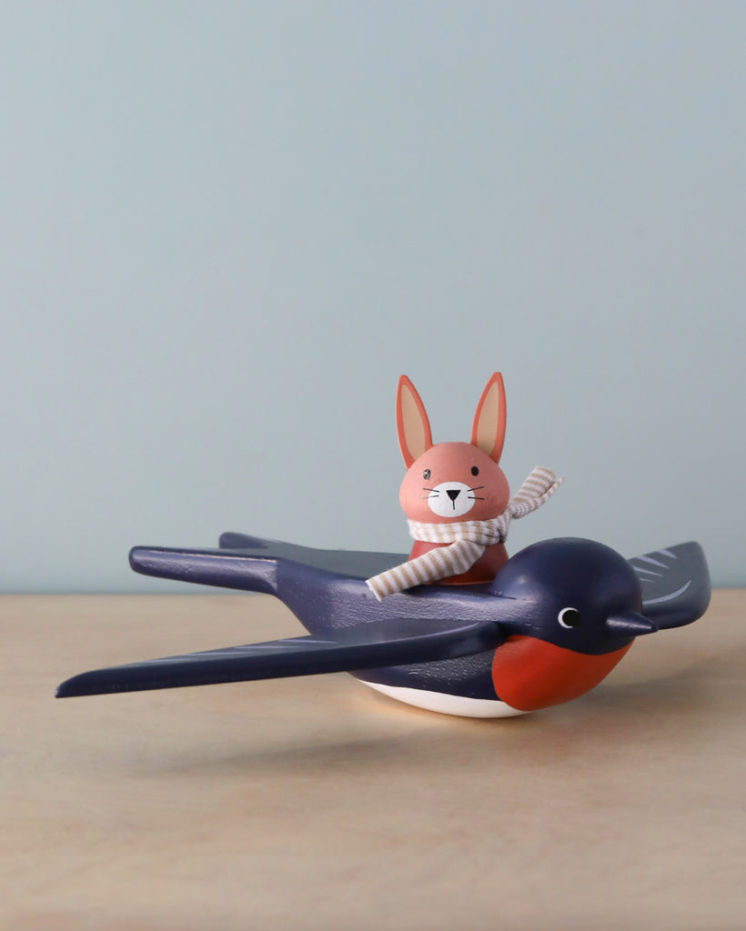 A toy rabbit named Spud the pilot Hare with a striped scarf, riding on Swifty Bird, a blue and red wooden bird figurine, set against a neutral beige background.