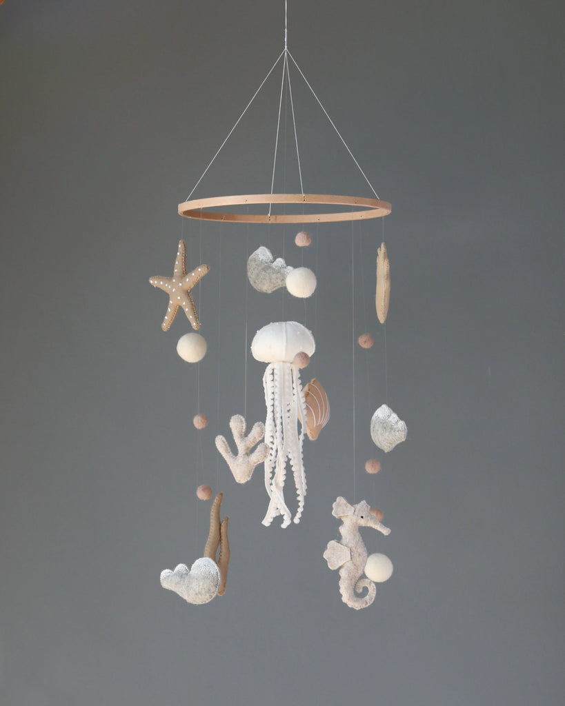 A Handmade Mobile - In The Ocean - Final Sale featuring a central white jellyfish surrounded by starfish, seashells, and coral, hanging from a circular frame against a gray background.