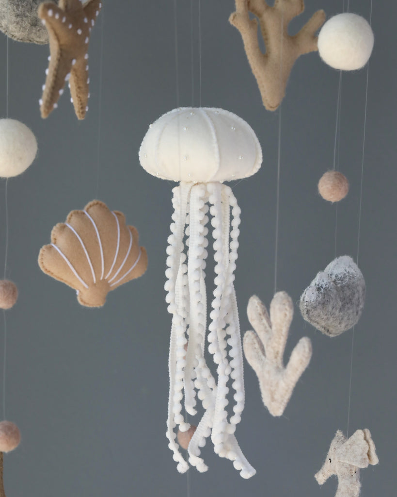 A nursery mobile featuring a Handmade Mobile - In The Ocean - Final Sale with flowing white tentacles, suspended alongside various felt sea creatures like shells and coral, set against a pale gray background.
