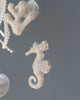A close-up of a felt seahorse ornament hanging by a string, part of the Handmade Mobile - In The Ocean - Final Sale , with soft-focus white beads in the background. Its delicate features and curled tail are highlighted against a
