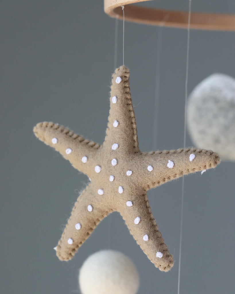 A close-up of a felt starfish ornament with white dots, hanging from the Handmade Mobile - In The Ocean - Final Sale alongside other spherical felt decorations, set against a soft gray background.