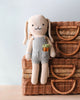 A Cuddle + Kind Bunny sitting on a wicker basket, holding a small knitted carrot. The rabbit has long floppy ears, a stitched smile, and is dressed in a gray outfit.