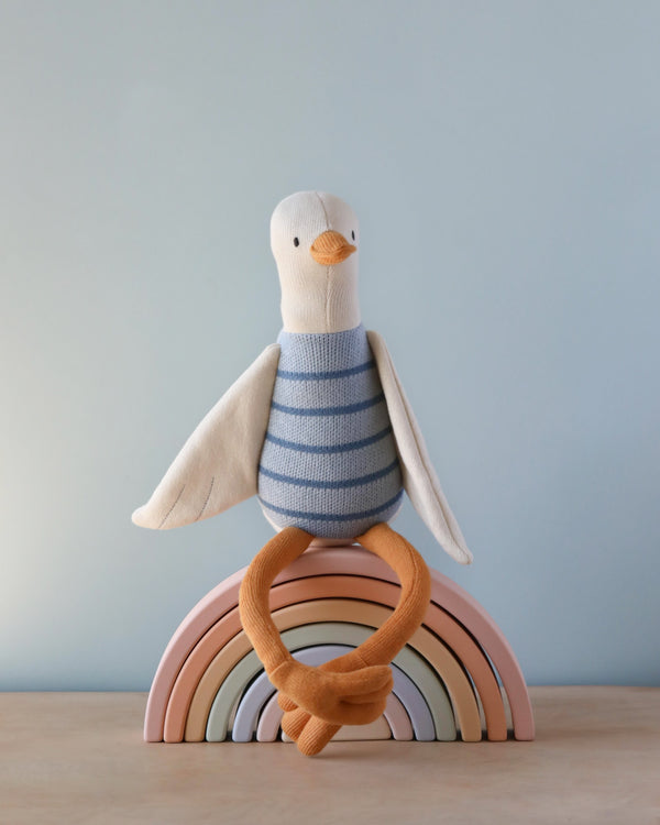 A Seagull Stuffed Animal with a striped, knitted organic cotton body and orange feet, perched playfully atop a wooden rainbow toy, set against a soft blue background.