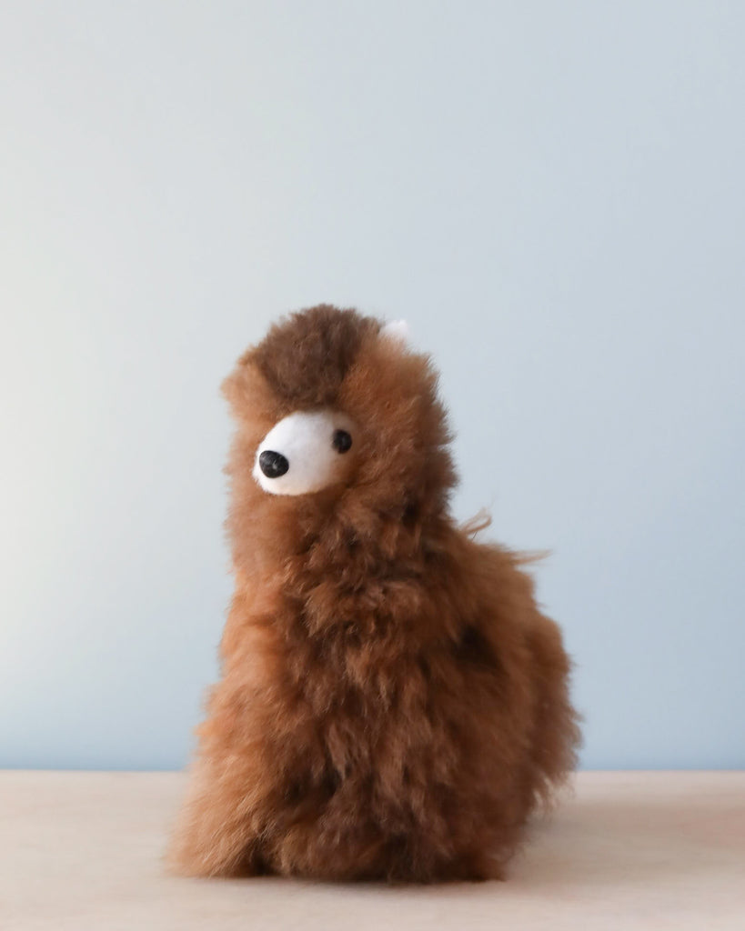 A Fluffy Alpaca Stuffed Animal - Small sits on a plain surface against a light blue background. The toy, crafted from handmade alpaca wool, features a round, white eye.