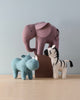 Three small, colorful, Olli Ella Holdie Folk Felt Safari Animals—a blue rhino, a pink elephant, and a white zebra with black stripes—arranged on a wooden surface against a plain, light background.