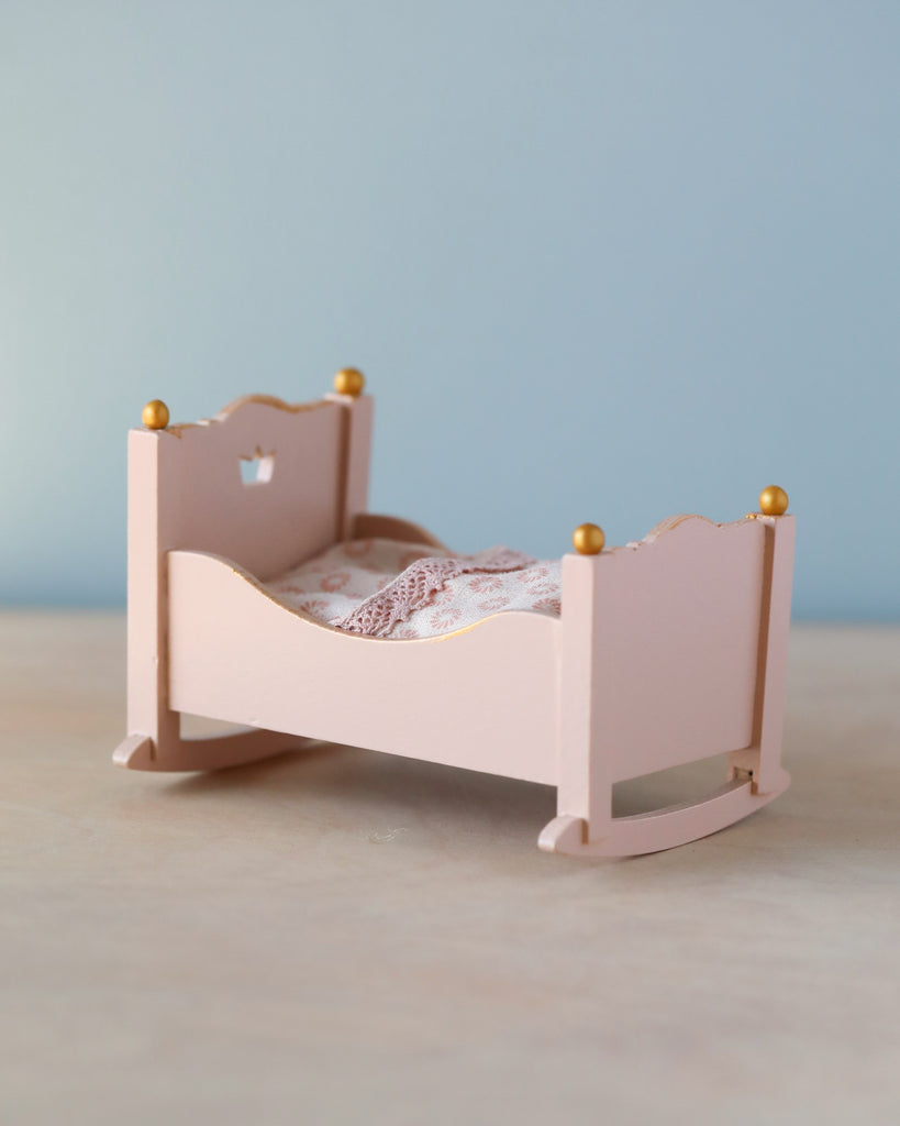 A Maileg Miniature Cradle with ornate details and golden knobs, set against a simple blue and beige background. The cradle is adorned with a delicate white lace bedding.