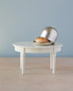 A single burger on a small Maileg Miniature Dining Table with a metal dome cover partially lifted, against a soft blue background.