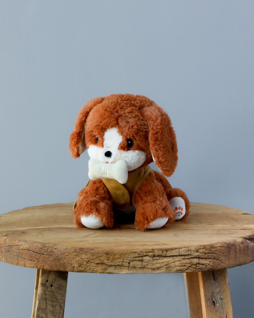 A plush toy of a Olli Ella Dinkum Dog with floppy ears, sitting on a round wooden stool against a plain gray background. The toy holds a magnetic bone accessory in its mouth.