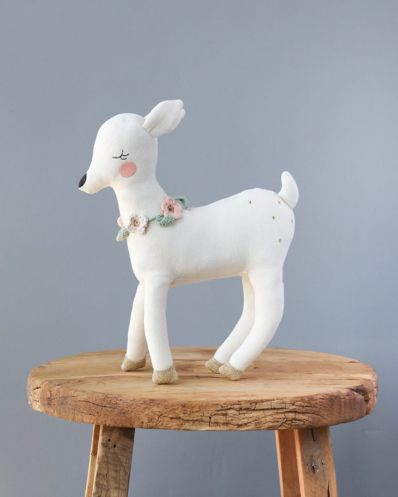 A Meri Meri Blossom Baby Deer Stuffed Animal with pink cheeks and closed eyes, adorned with a crochet flower necklace around its neck, stands on a wooden stool against a gray background.