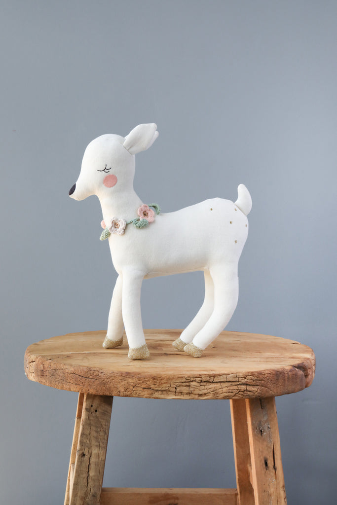 A Meri Meri Blossom Baby Deer Stuffed Animal with a crochet flower necklace, standing gracefully on a round wooden stool against a plain gray background.