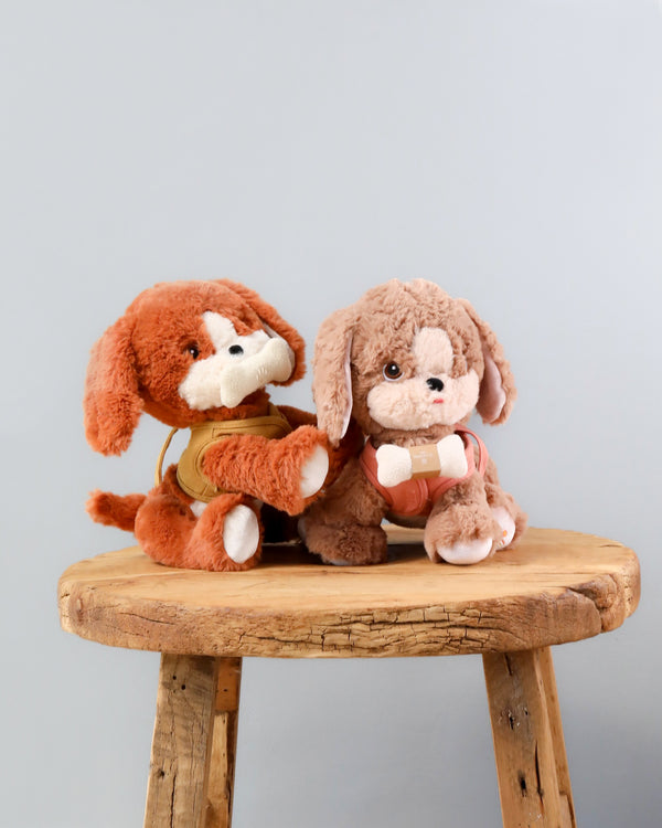 Two Olli Ella Dinkum Dogs, one orange and one light brown, sitting on a wooden stool against a gray background. Crafted from plush fabric, the toys appear soft and cuddly, with distinct floppy ears.