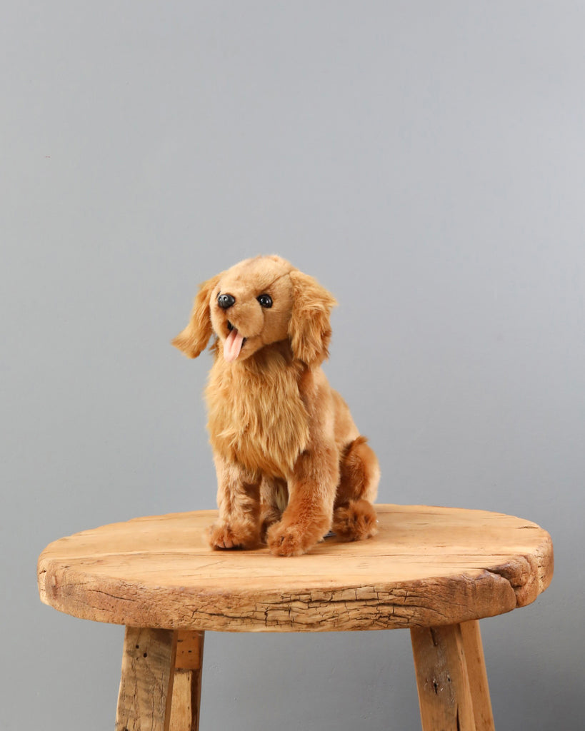 A Golden Retriever Dog Stuffed Animal with light brown fur sitting on a circular wooden stool against a grey background. The hand-sewn plush dog has a perky expression with its tongue out.