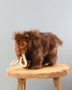 A high-quality Mammoth Stuffed Animal resembling a woolly mammoth with brown fur and prominent white tusks, displayed on a round wooden stool against a plain grey background.