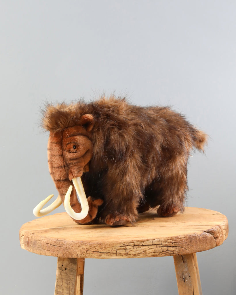 A high-quality Mammoth Stuffed Animal resembling a woolly mammoth with brown fur and prominent white tusks, displayed on a round wooden stool against a plain grey background.