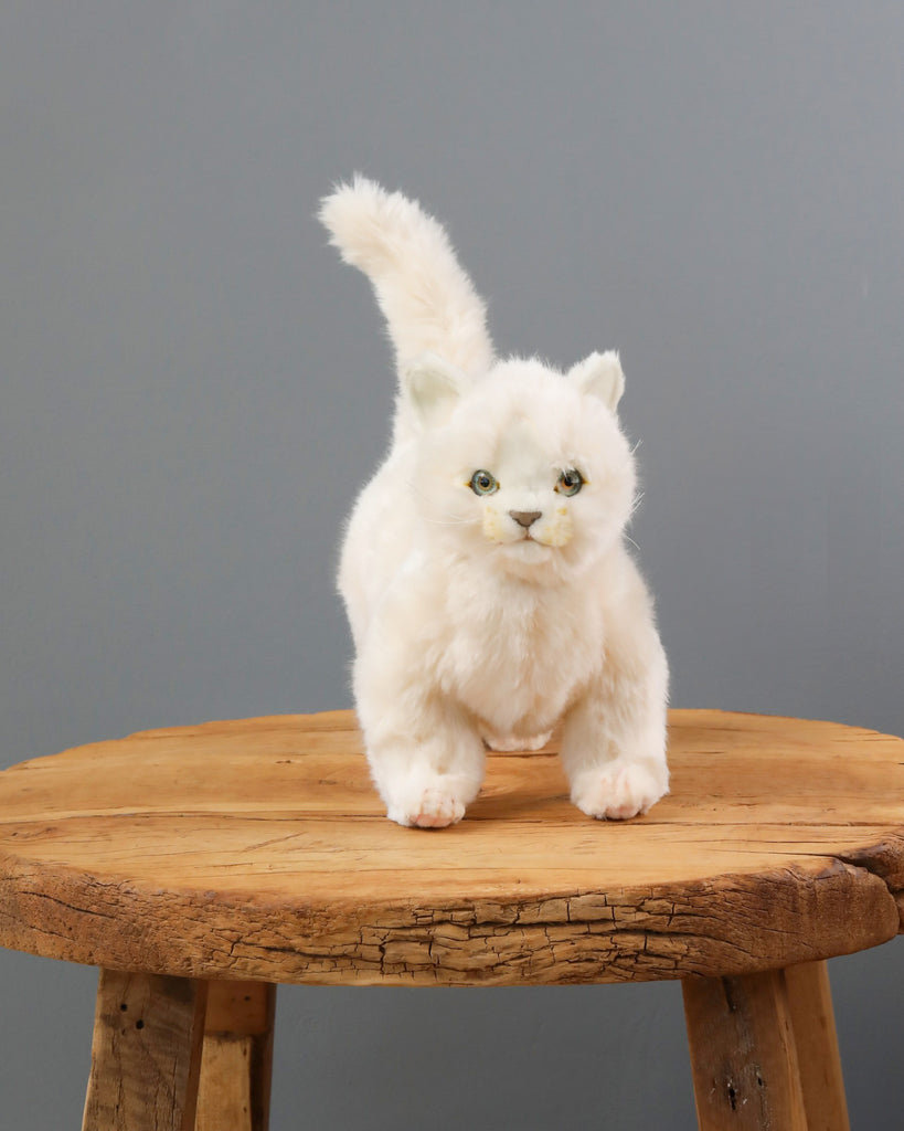 A White Cat Stuffed Animal with bright yellow eyes, positioned on a round wooden table against a solid grey background. The stuffed animal has realistic plush features and a fluffy tail pointed upwards.