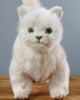 A lifelike White Cat Stuffed Animal with vivid green eyes standing on a wooden table against a gray background. The toy, hand-sewn with realistic plush features, has delicate facial features.