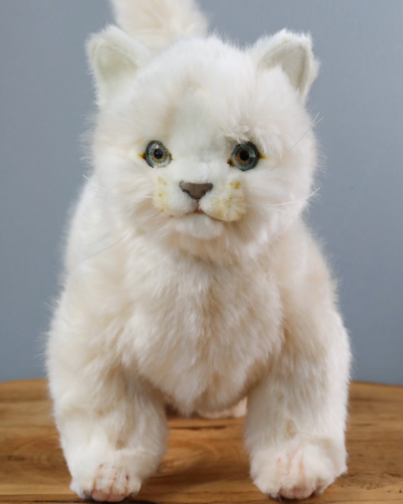 A lifelike White Cat Stuffed Animal with vivid green eyes standing on a wooden table against a gray background. The toy, hand-sewn with realistic plush features, has delicate facial features.