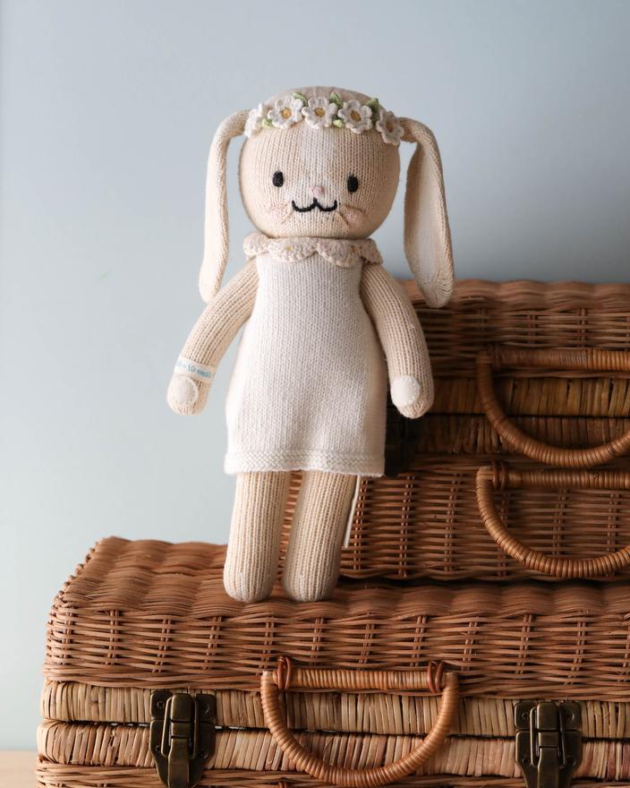 A handmade knitted Cuddle + Kind Bunny filled with hypoallergenic polyfill, dressed in a white dress and floral headband, stands on a wicker basket with a neutral background.
