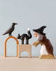 Decorative ceramic figurines depicting The Witch and Her Cat & Crow, arranged with a wooden arch on a table against a plain, light blue background.