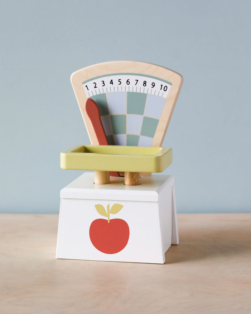 A child's Market Scale for pretend play shopping, with a pastel-colored weight indicator marked from 1 to 10, set on a plain white block decorated with a red apple design, against