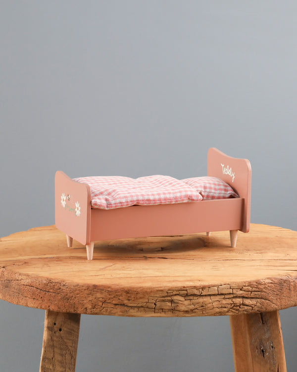A small pink Wooden Bed for Teddy Mum - Rose with plaid bedding and handpainted details on it, labeled "Teddy" and decorated with white snowflakes, placed centrally on a rustic wooden table against a plain