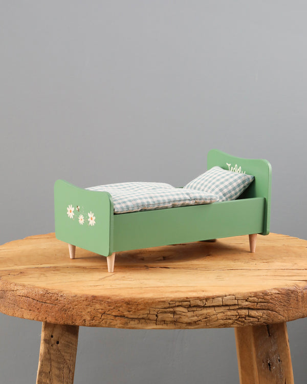 A small Wooden Bed for Teddy Dad - Dusty Green with a checkered mattress placed on a rustic wooden table against a gray background. The bed features handpainted floral designs on the sides.