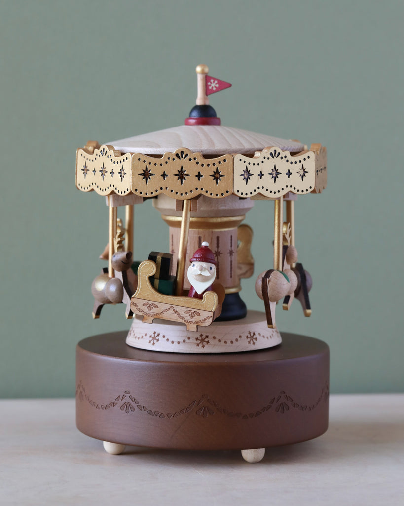 A Christmas Carousel Music Box with ornate designs. It features miniature horses and chariots, with a Santa Claus figure in one of them. Crafted from sustainably sourced wood, the base is brown with intricate patterns. A festive flag tops the carousel against a green background.