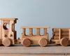 A Handmade Wooden Train - Extra Long, composed of a locomotive and two cars on a beech wood surface against a light blue background. The train features circular wheels and cut-out windows.
