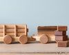 A handmade wooden train with a detachable flatbed trailer, carrying square blocks, against a plain, light blue background.