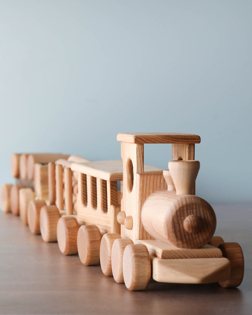 A Handmade Wooden Train - Extra Long set consisting of an engine and two carriages, displayed on a table against a soft blue background. Each component is crafted with visible wheels and details.