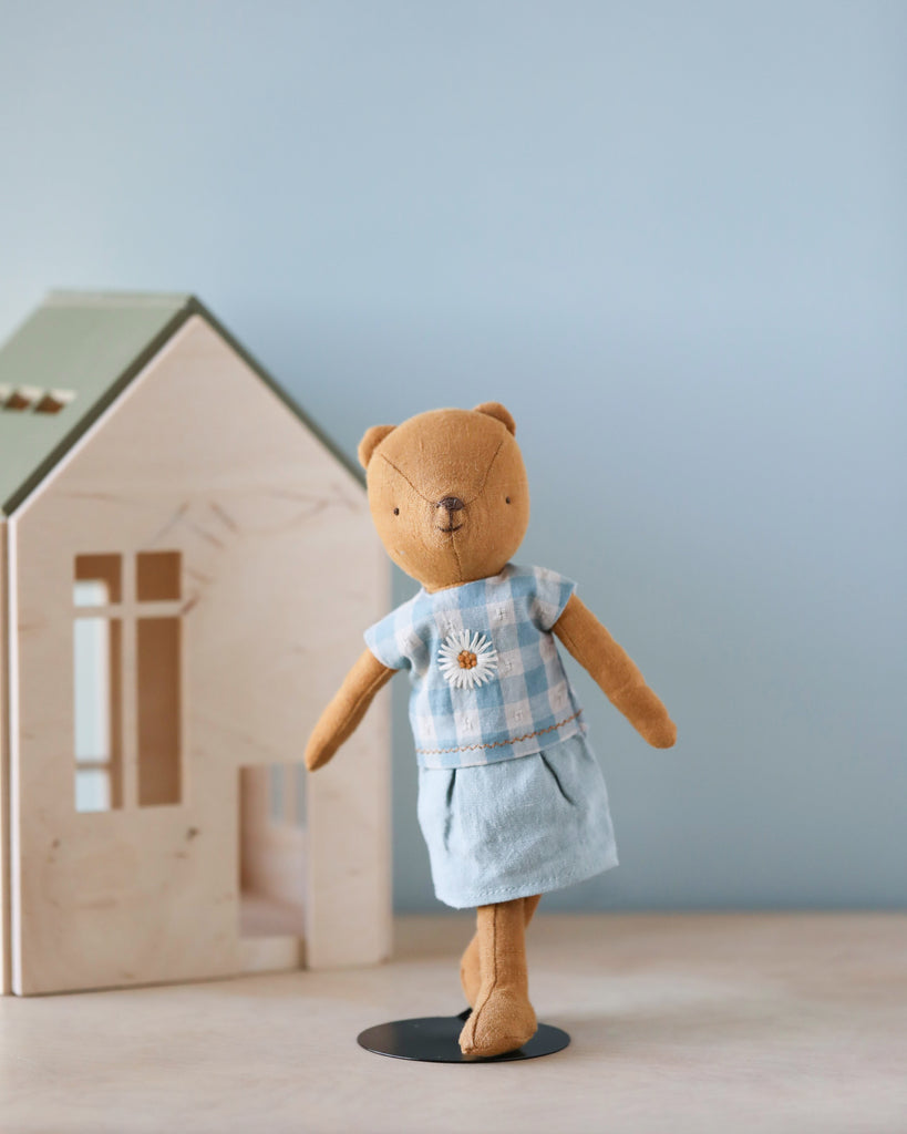 A Maileg Extra Clothing: Dress for Teddy Mum in a light blue outfit with a flower detail stands on one leg in front of a wooden toy house with a green roof. The background is a soft blue wall.