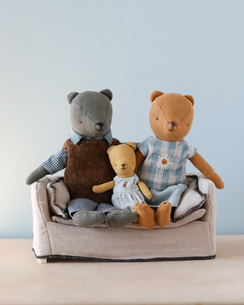 Three plush teddy bears—a gray one, a tan one, and a small yellow one—sit closely together on a Maileg Dollhouse Couch against a light blue background.