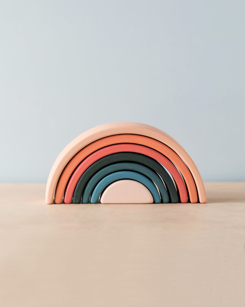 A Handmade Mini Rainbow Stacker - Tropics consisting of seven arcs in shades of pink, orange, and blue, stacked progressively on a light wooden surface against a pale blue background.